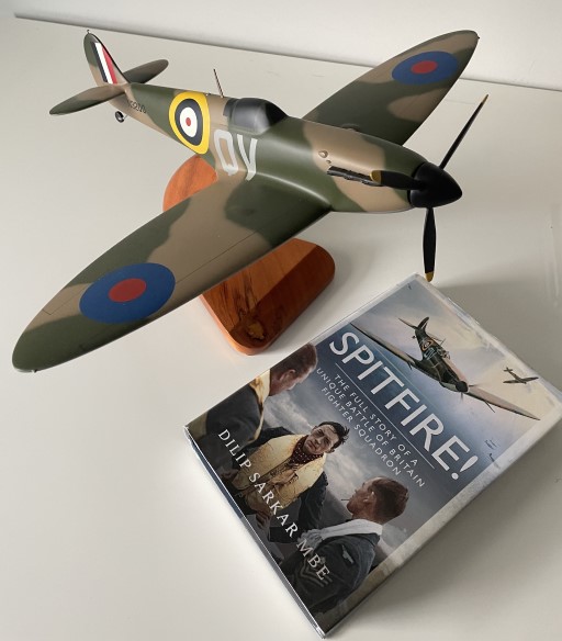 Spitfire Mk I model with camouflage paint, RAF roundels, and code QY on wooden stand beside Spitfire history book