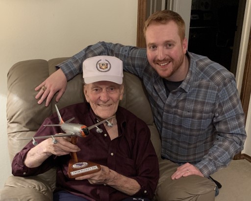 Two people smiling indoors, one seated holding Marine aircraft model on wooden stand with plaque reading CMSgt Richard S. Doll.