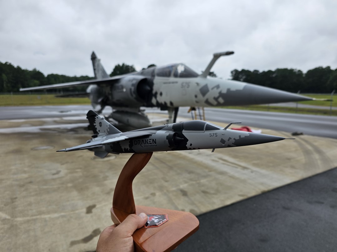 Mirage F1 model with gray digital camouflage and “DRAKEN 575” held in front of a matching real jet
