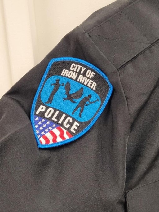 City of Iron River Police embroidered shoulder patch with blue border, symbolic figures, and U.S. flag detail on black uniform sleeve.