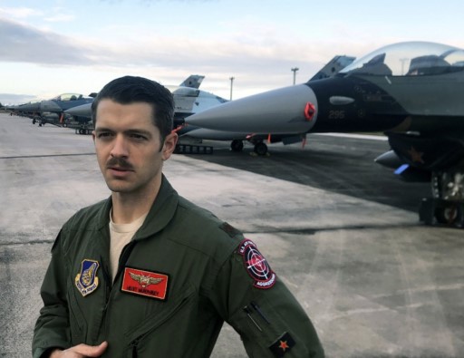 An airman in a flight suit standing on an airfield with fighter jets in the background