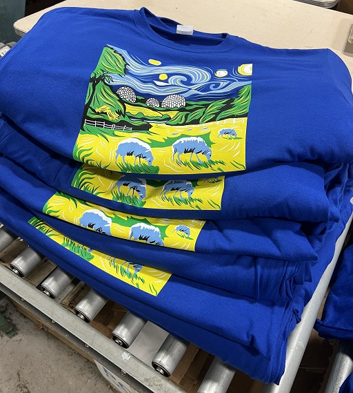 Stack of RAF Menwith Hill shirts with colorful printed front design showing hills, sheep, and fence